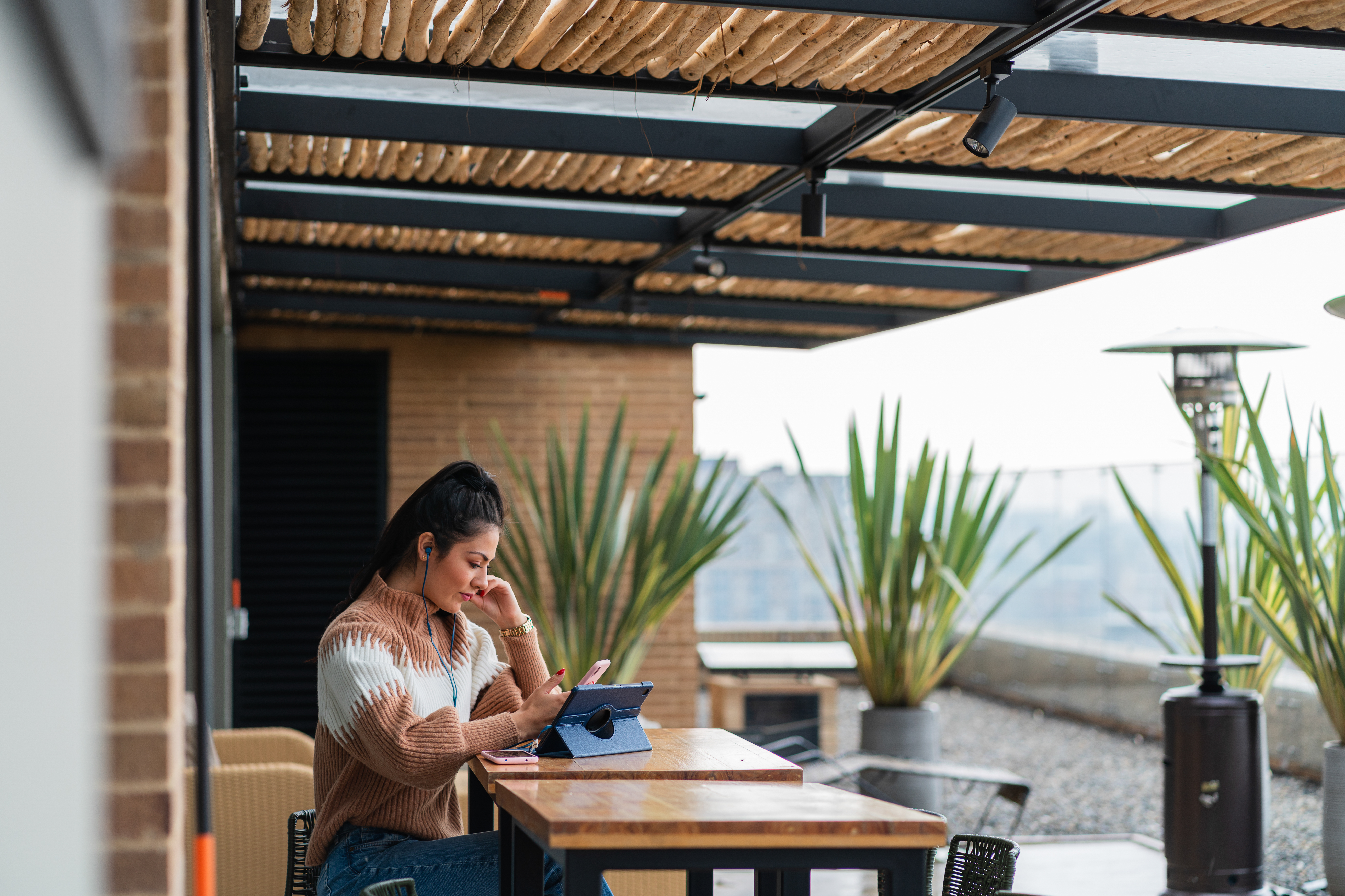 Latin woman of approximately 25 years dressed in an informal way is sitting on the terrace of a shopping center with her headphones in her ears which are connected to her digital tablet through which she interacts virtually