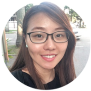 A smiling woman with long hair and glasses, wearing a black shirt, is pictured outdoors on a street with trees and cars in the background.